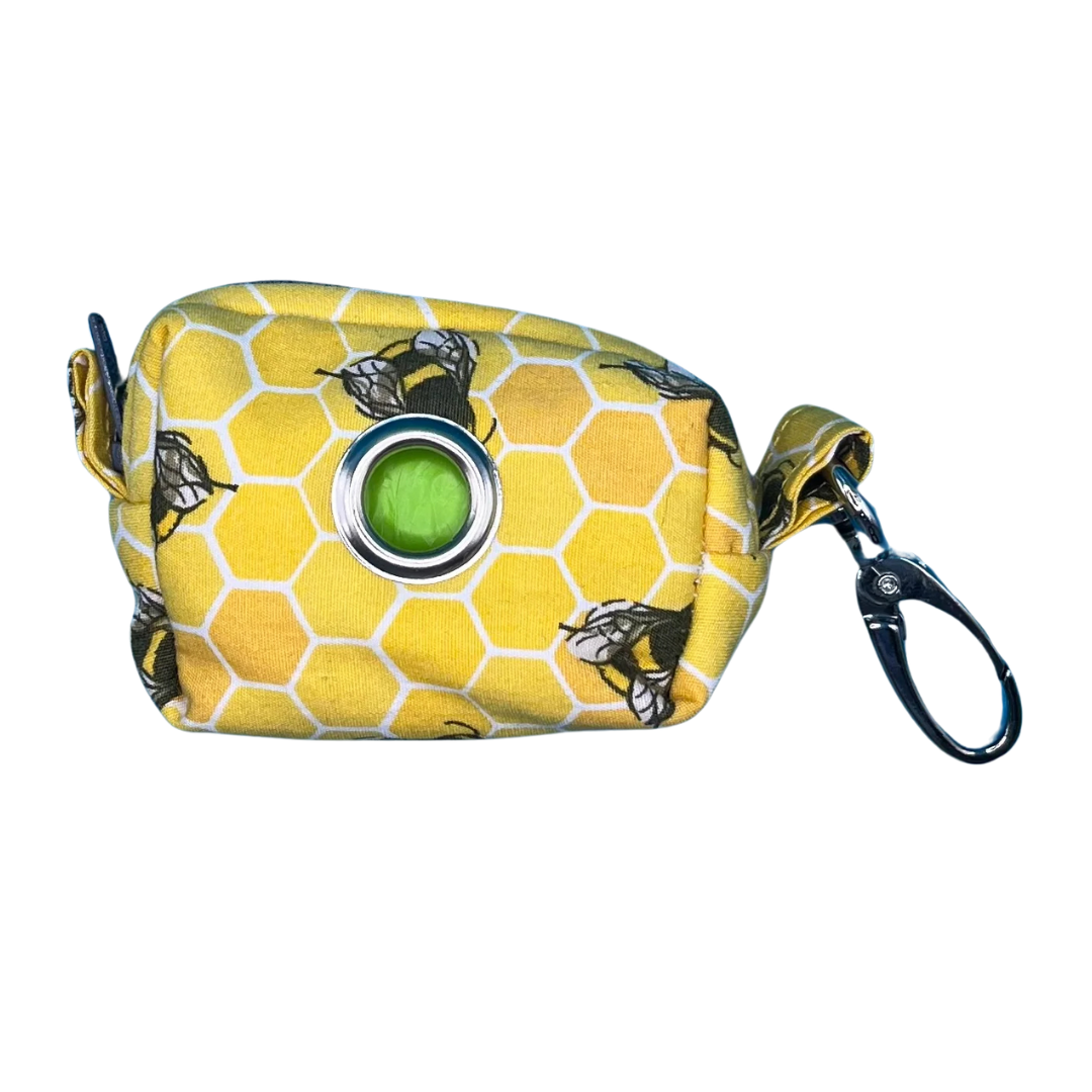 A fabric bee hive poo bag holder with a yellow honeycomb pattern and black bees, featuring a black zip and metal accessories.