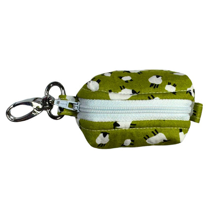 Top view of a fabric poo bag holder with a green background and white sheep patterns, featuring a white zip and a metal keychain.