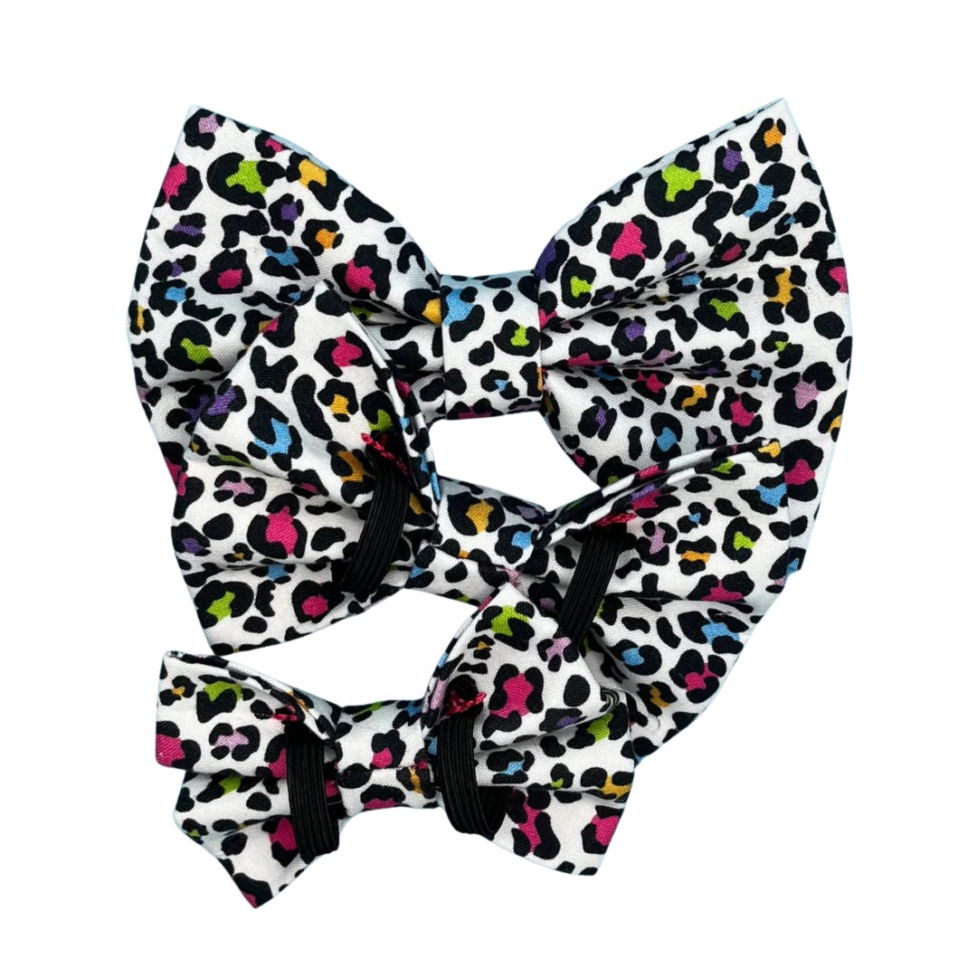 A multicoloured leopard print dog bow tie with a white background in three sizes, small, medium and large showing the double elastic.