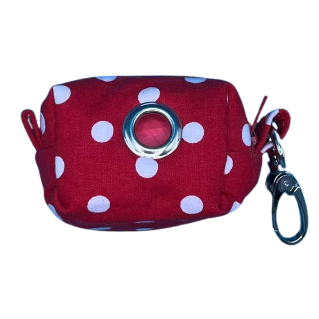 A red fabric poo bag holder with white polka dots a metal clip. It also features a roll of compostable poo bags.