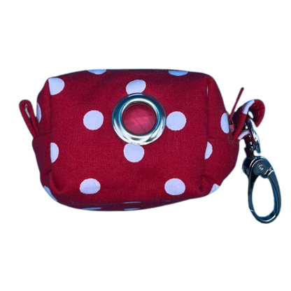 A red fabric poo bag holder with white polka dots a metal clip. It also features a roll of compostable poo bags.