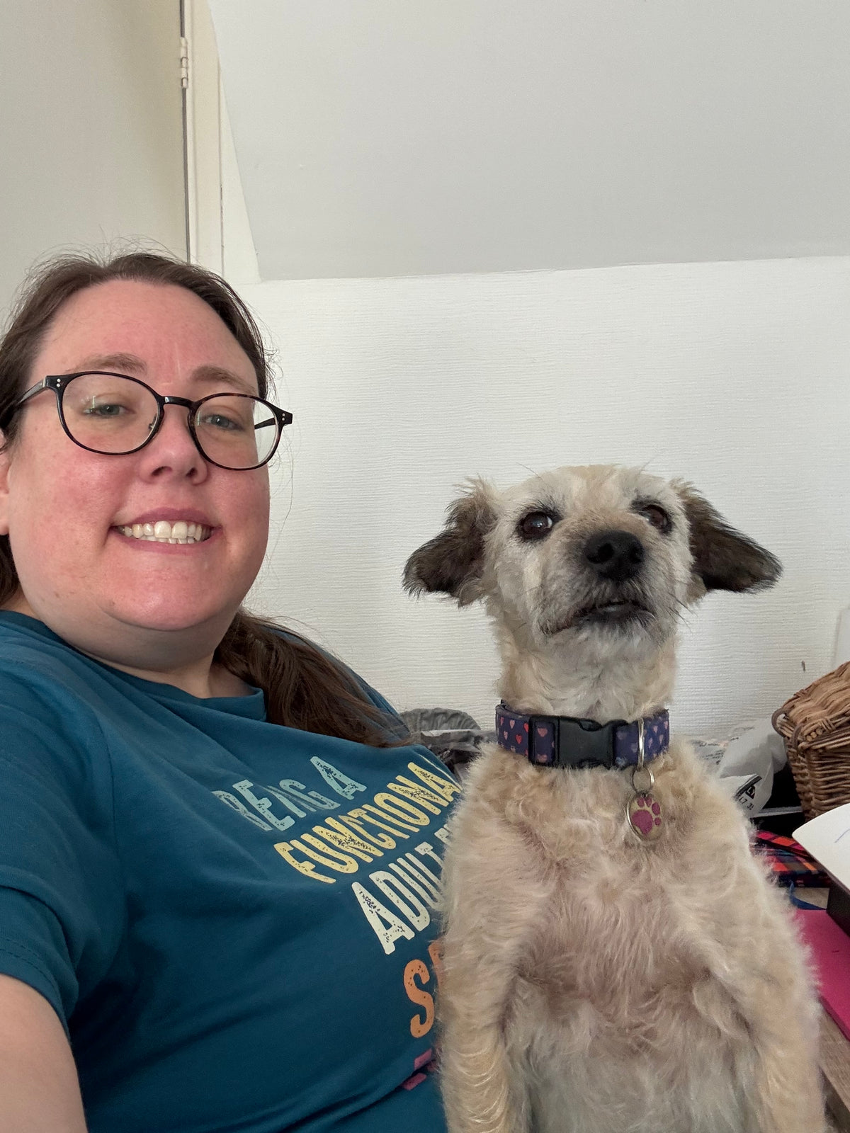 A woman wearing a blue graphic t-shirt and glasses smiles next to a small scruffy light-colored dog with a purple patterned collar, in an indoor setting.