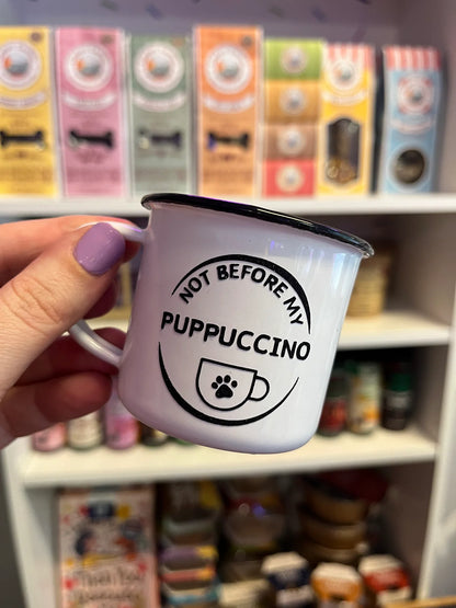 A white enamel cup with a 'Not Before my Puppuccino' decal held in a person's hand, with various food and drink items in the background.