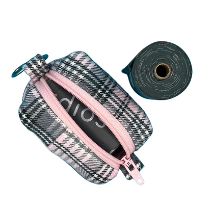 A baby pink and grey tartan patterned fabric poo bag holder with a pink zipper that is open, metal accessories, alongside a roll of black poo bags.