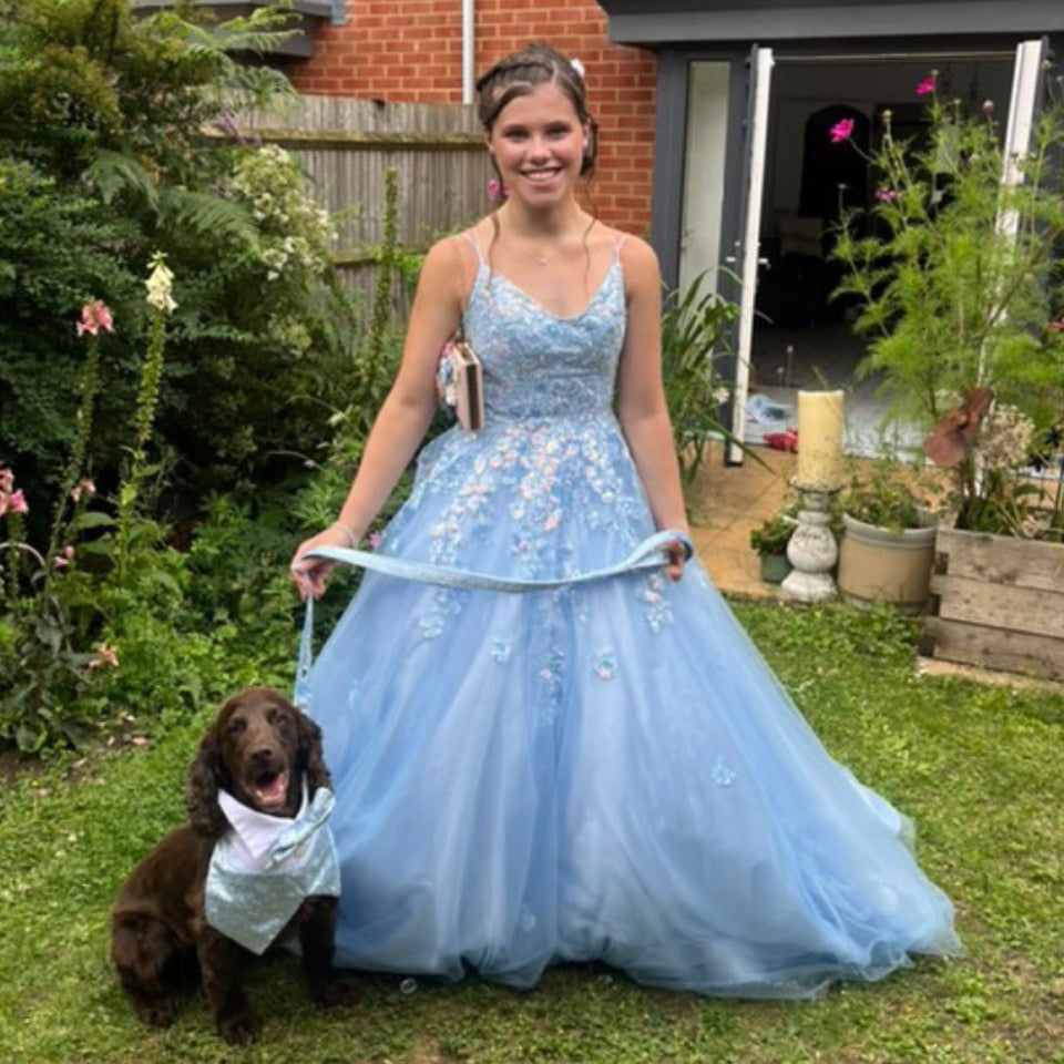 Ready for prom, person in a blue formal gown standing with a dog wearing a waistcoat and bow tie in a garden.