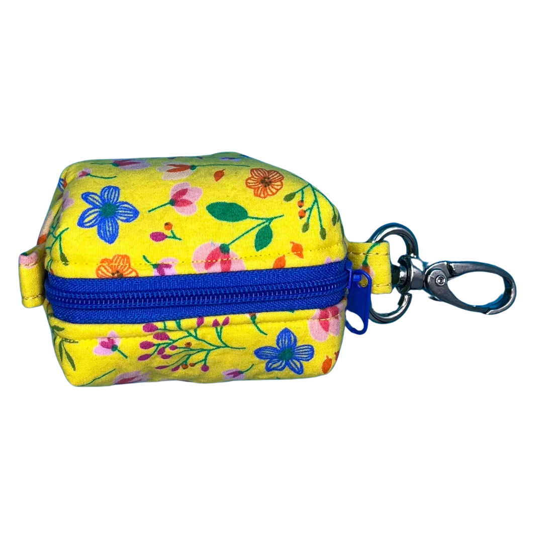 A top view of a yellow fabric poo bag holder with a floral pattern, featuring a metal clip and a blue zipper.